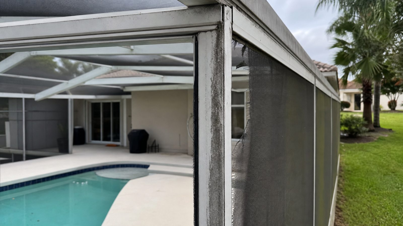 Florida pool cage corner with torn mesh along spline channel, oxidized aluminum frame, afternoon light